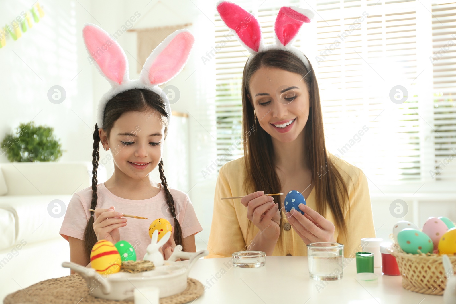 Photo of Happy mother and daughter with bunny ears headbands painting Easter eggs at home