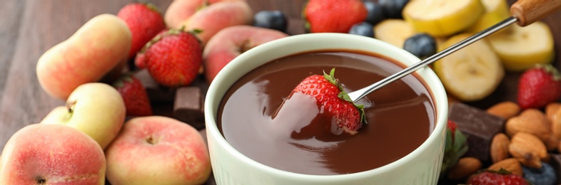 Fondue fork with strawberry in bowl of melted chocolate surrounded by other fruits on wooden table