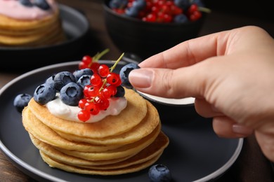 Photo of Woman decorating tasty pancakes with natural yogurt, blueberries and red currants at wooden table, closeup