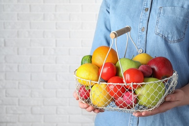 Photo of Woman holding basket with ripe fruits and vegetables near white brick wall, closeup. Space for text