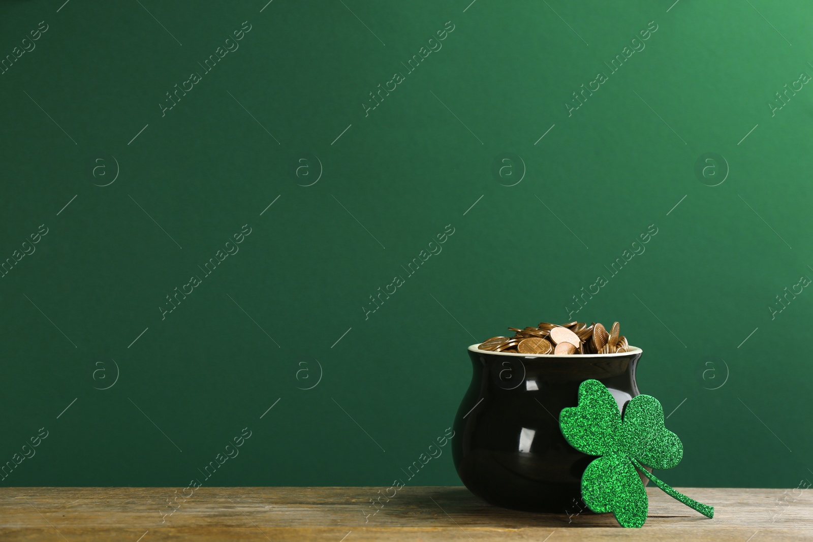 Photo of Pot of gold coins and clover on wooden table against green background, space for text. St. Patrick's Day celebration