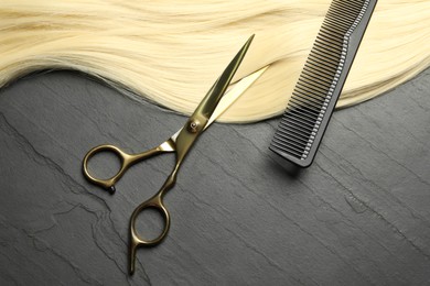 Professional hairdresser scissors and comb with blonde hair strand on dark grey table, flat lay