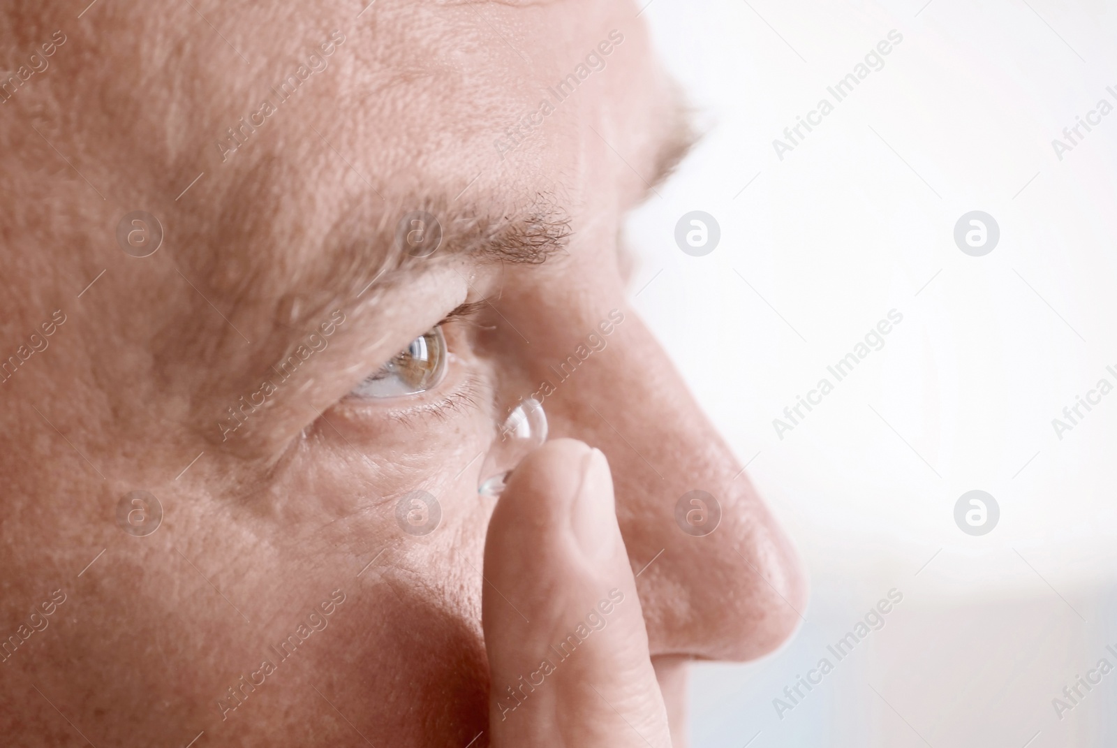 Photo of Mature man putting contact lens in his eye on light background, closeup