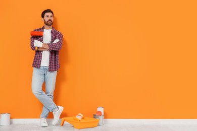 Photo of Designer with roller and painting equipment on floor near freshly painted orange wall indoors, space for text
