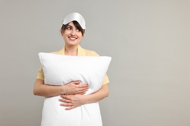 Photo of Happy woman in pyjama and sleep mask holding pillow on grey background, space for text