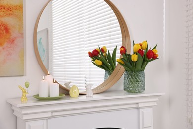 Photo of Easter decorations. Bouquet of tulips in vase, burning candles and bunny figures on fireplace at home