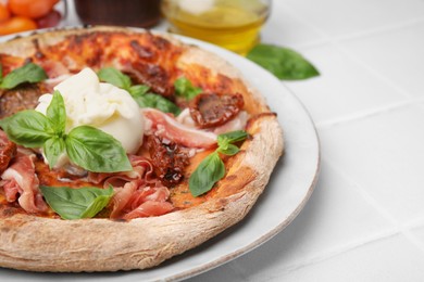 Delicious pizza with burrata cheese, basil, ham and sun-dried tomatoes on white tiled table, closeup. Space for text