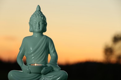 Decorative Buddha statue with burning candle outdoors at sunset. Space for text