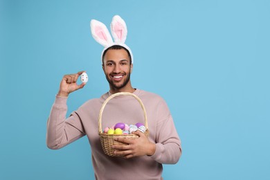 Happy African American man in bunny ears headband holding wicker basket with Easter eggs on light blue background. Space for text