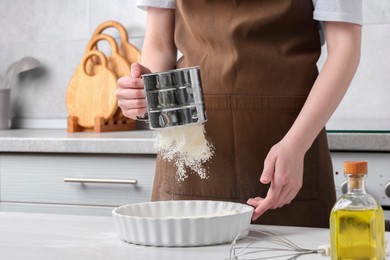 Photo of Woman sieving flour into baking dish at table in kitchen, closeup