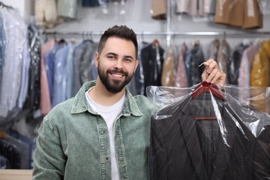 Photo of Dry-cleaning service. Happy man holding hanger with jacket in plastic bag indoors
