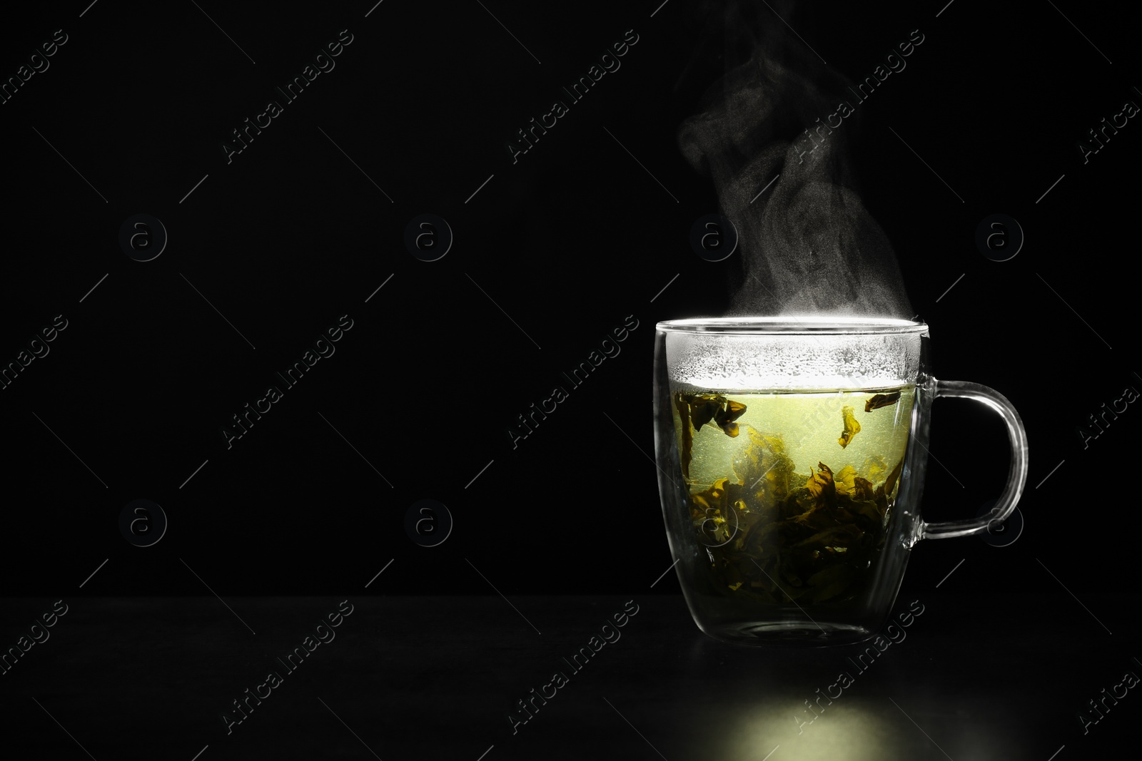 Photo of Glass cup of hot green tea on table against black background, space for text