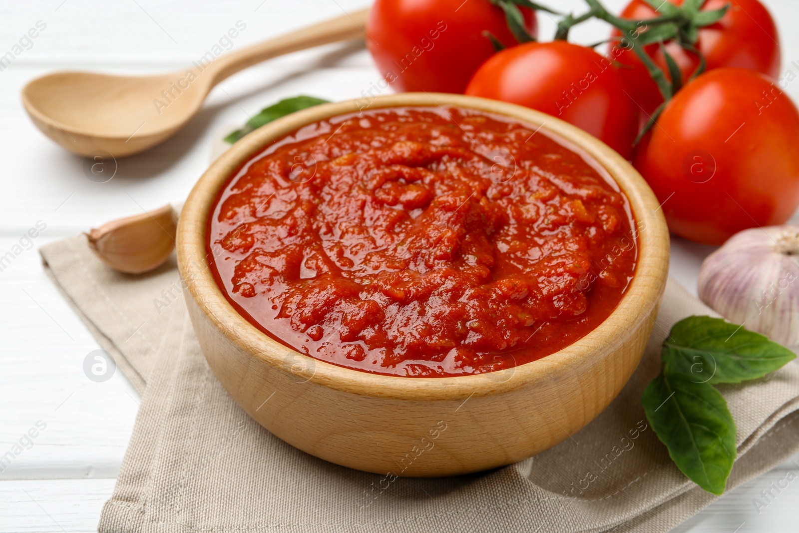 Photo of Homemade tomato sauce in bowl, spoon and fresh ingredients on white wooden table, closeup