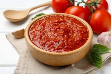 Photo of Homemade tomato sauce in bowl, spoon and fresh ingredients on white wooden table, closeup
