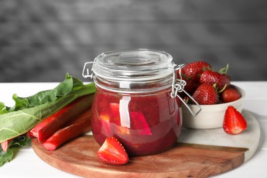 Photo of Jar of tasty rhubarb jam, fresh stems and strawberries on white table