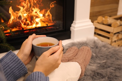 Woman with cup of tea sitting near burning fireplace, closeup