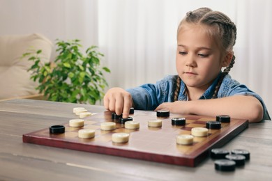 Photo of Playing checkers. Little girl thinking about next move at table in room
