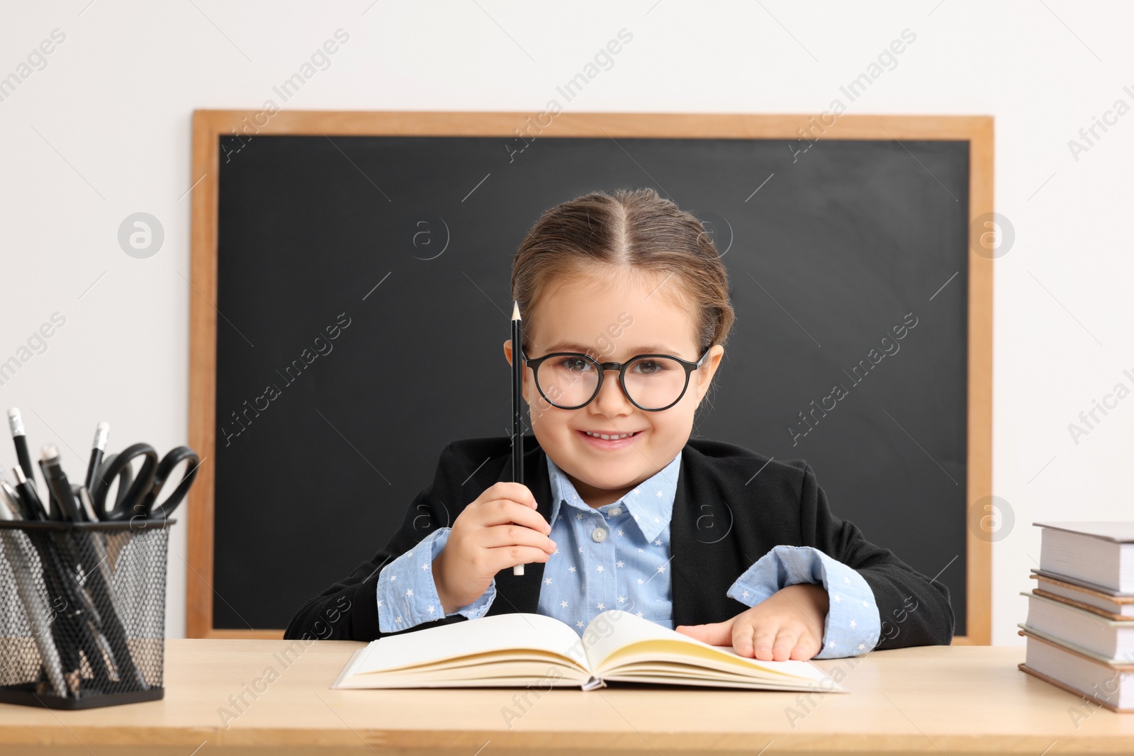 Photo of Happy little school child sitting at desk with books near chalkboard in classroom