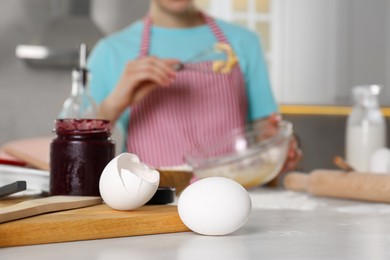 Photo of Woman cooking at messy countertop in kitchen, selective focus. Jar of jam, dishware, eggshells and utensils on table