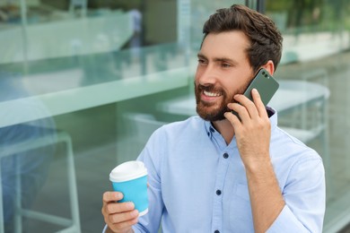Handsome man with cup of coffee talking on phone outdoors