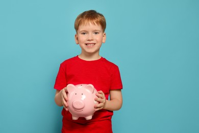 Cute little boy with ceramic piggy bank on light blue background