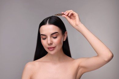 Beautiful young woman using ampoule for hair treatment on grey background