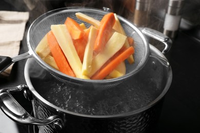 Sieve with cut parsnips and carrots over pot of boiling water in kitchen, closeup