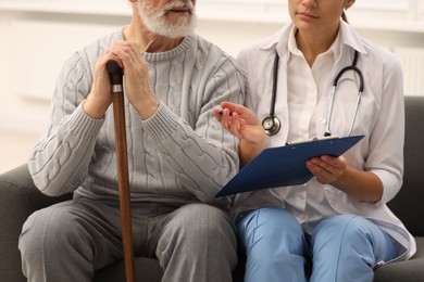 Photo of Nurse with clipboard assisting elderly patient on sofa indoors, closeup