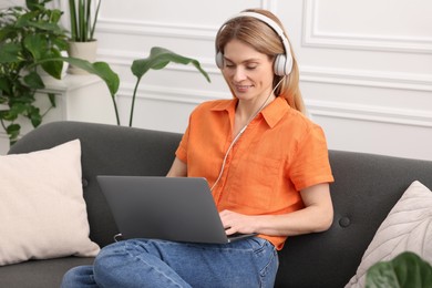 Woman in headphones using laptop on sofa near beautiful potted houseplants at home