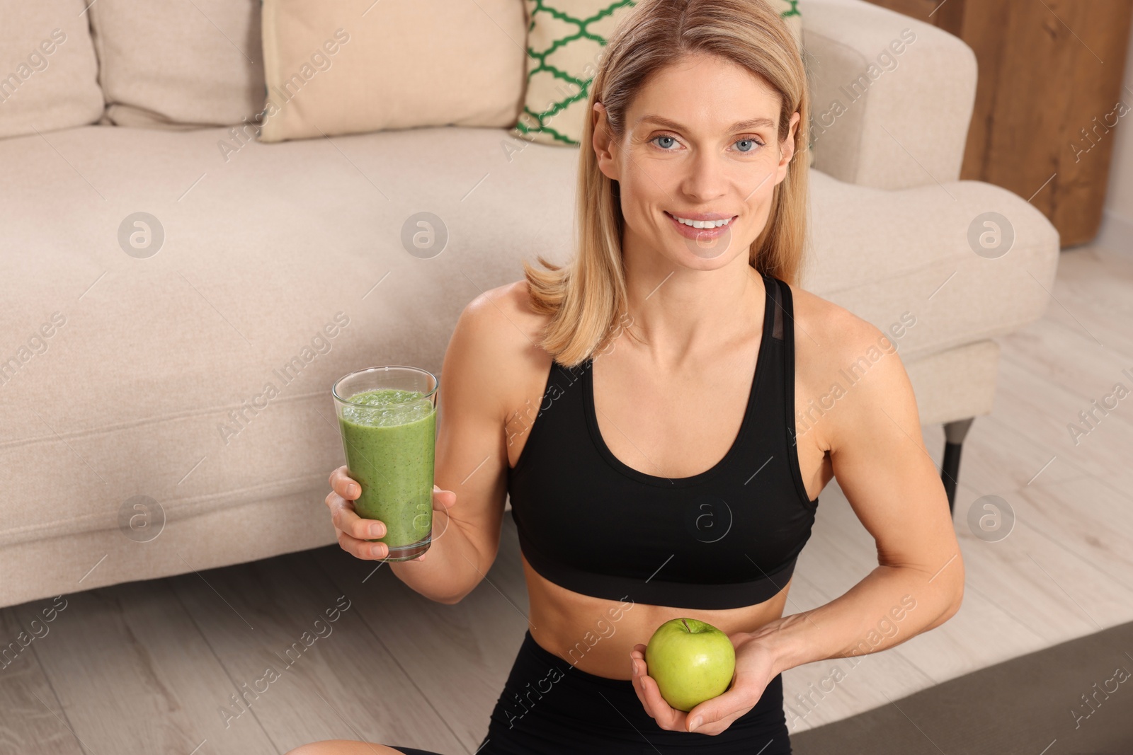 Photo of Young woman in sportswear with glass of fresh smoothie and apple at home