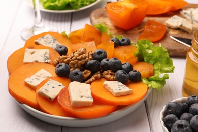 Delicious persimmon with blue cheese, blueberries, lettuce and nuts on white wooden table, closeup