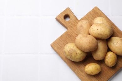 Photo of Raw fresh potatoes and wooden board on white tiled table, top view. Space for text