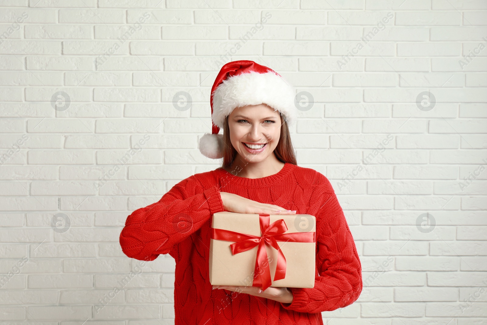 Photo of Happy young woman in Santa hat with Christmas gift near white brick wall