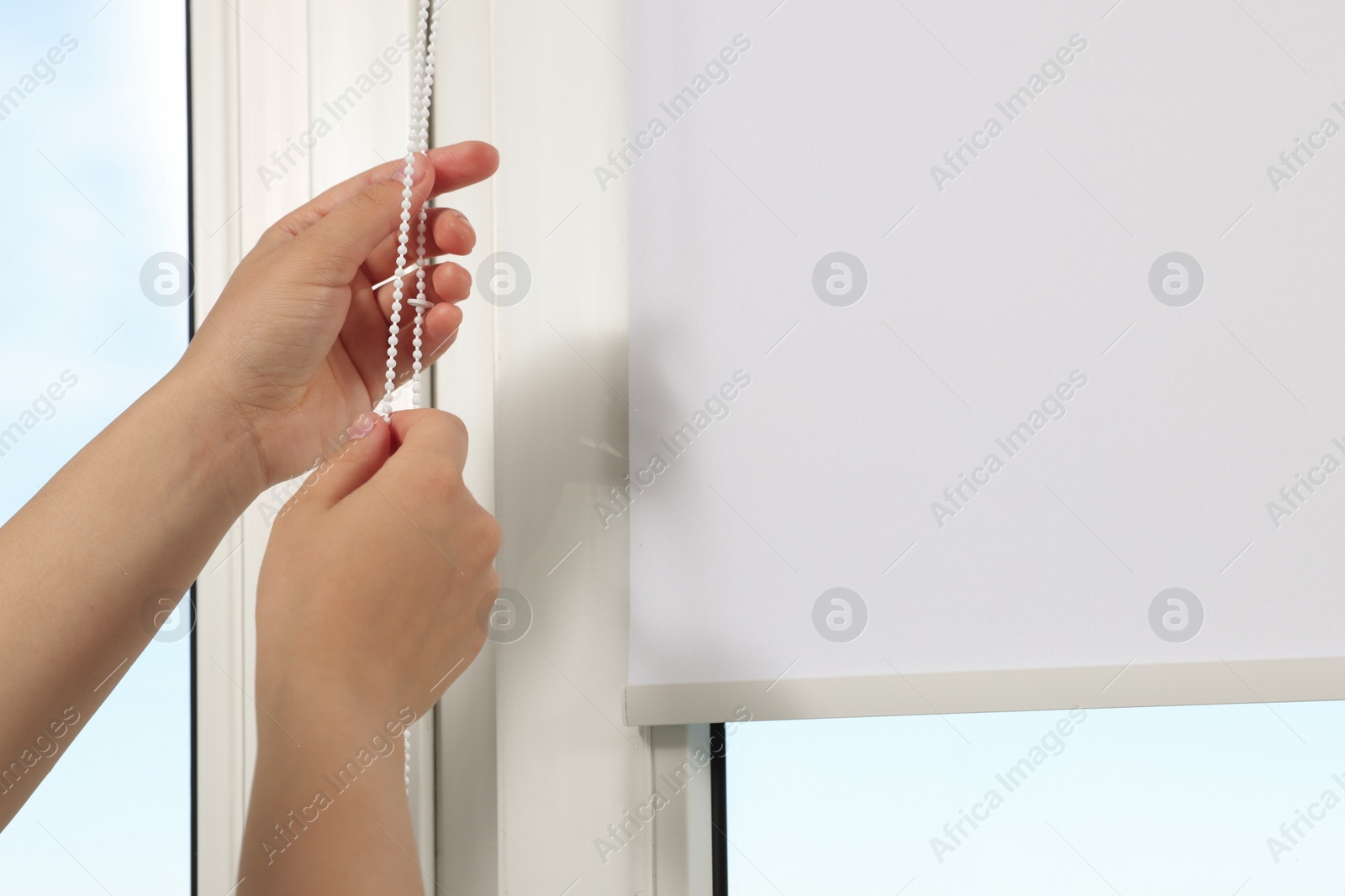 Photo of Woman opening white roller blind on window indoors, closeup