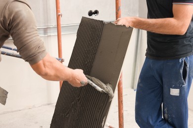 Photo of Worker spreading adhesive mix over tile with spatula, closeup