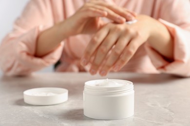 Photo of Woman applying cosmetic cream onto hand at grey table, focus on jar