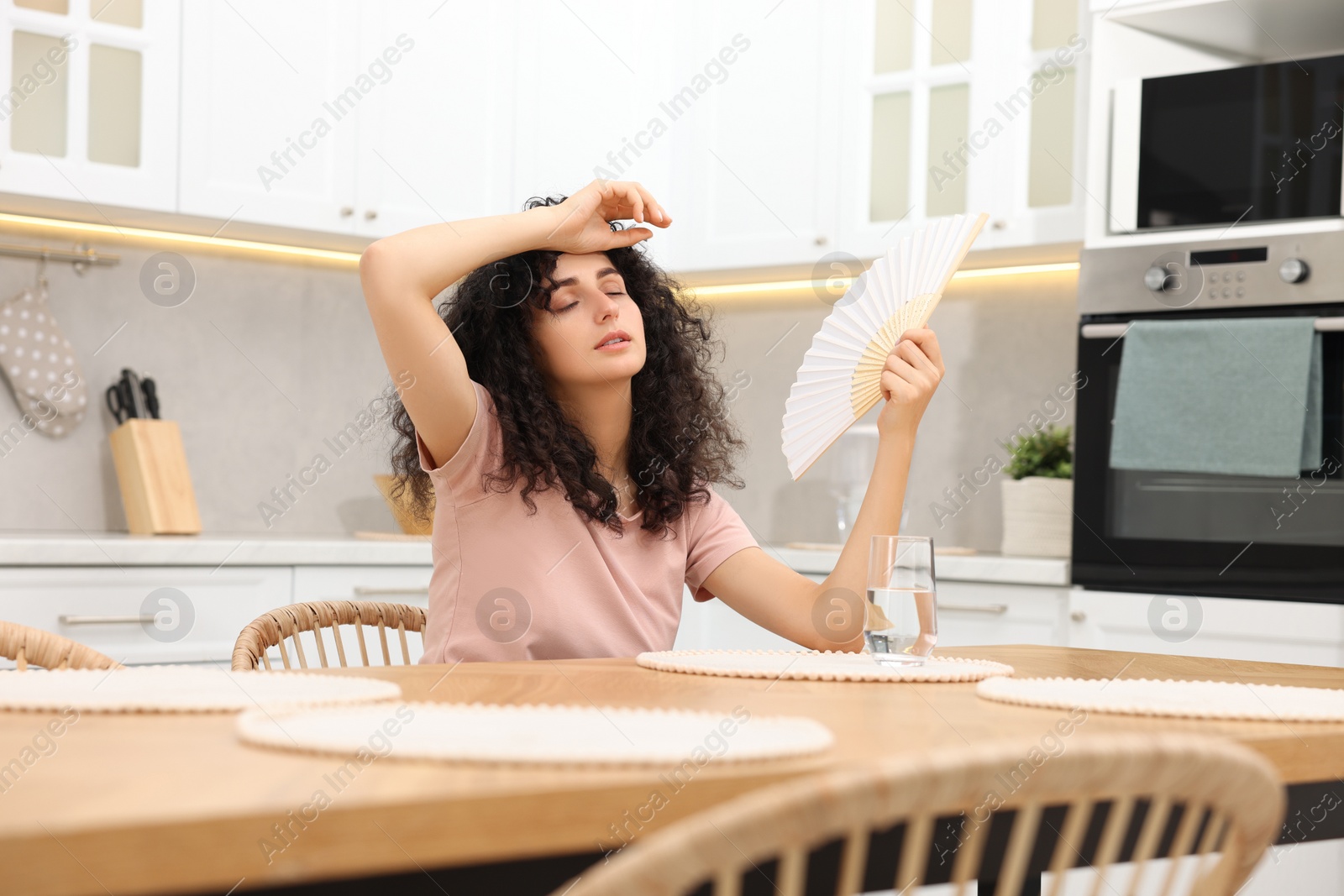 Photo of Young woman waving hand fan to cool herself at table in kitchen