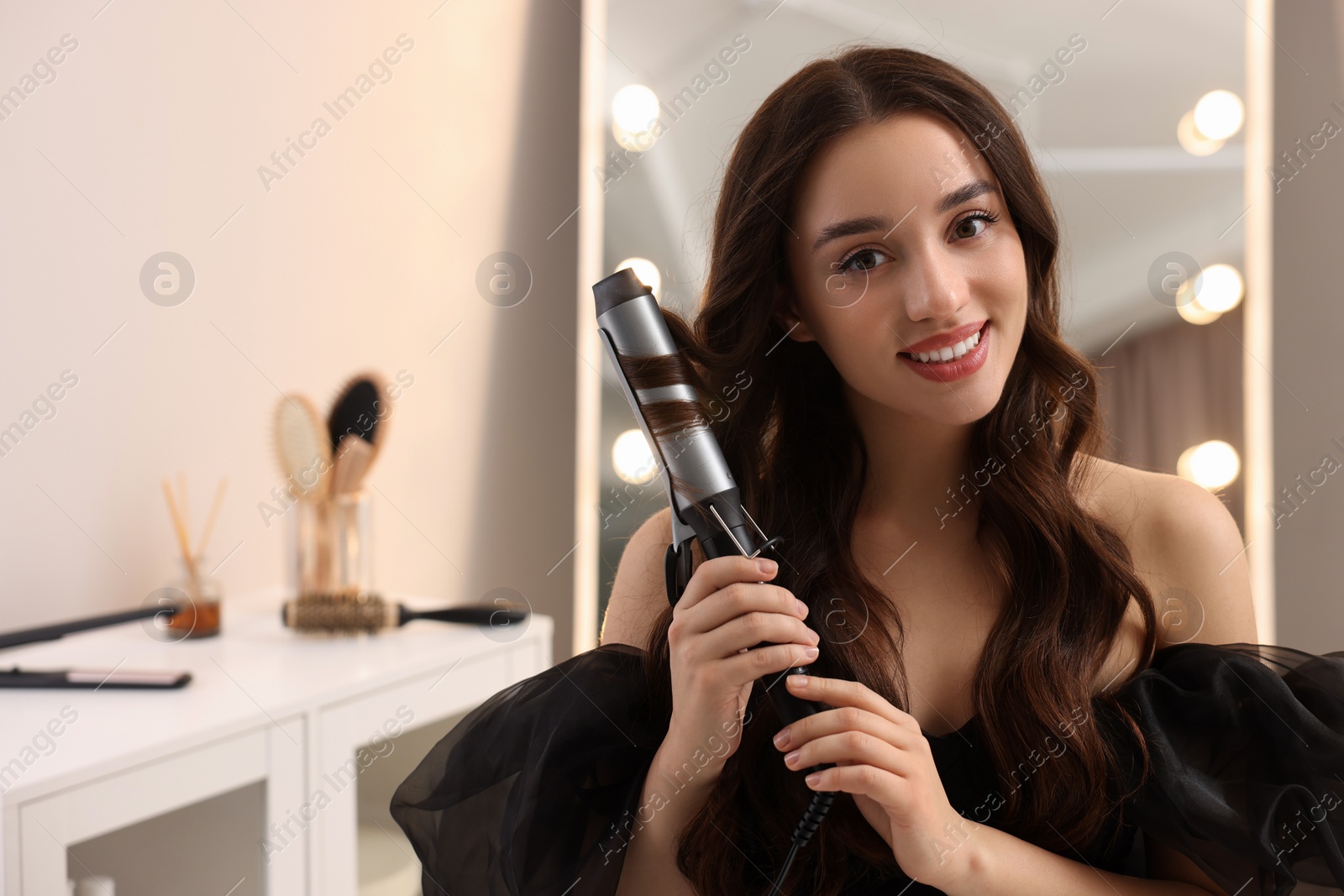 Photo of Smiling woman using curling hair iron at home. Space for text