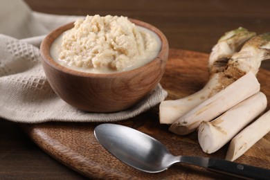 Photo of Spicy horseradish sauce in bowl, roots and spoon on wooden table, closeup