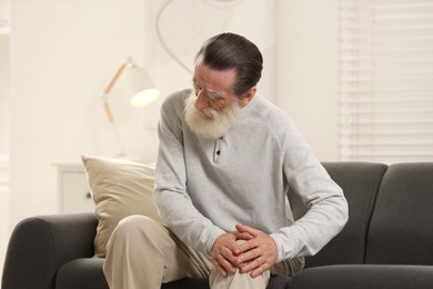 Photo of Senior man suffering from knee pain on sofa at home. Rheumatism symptom