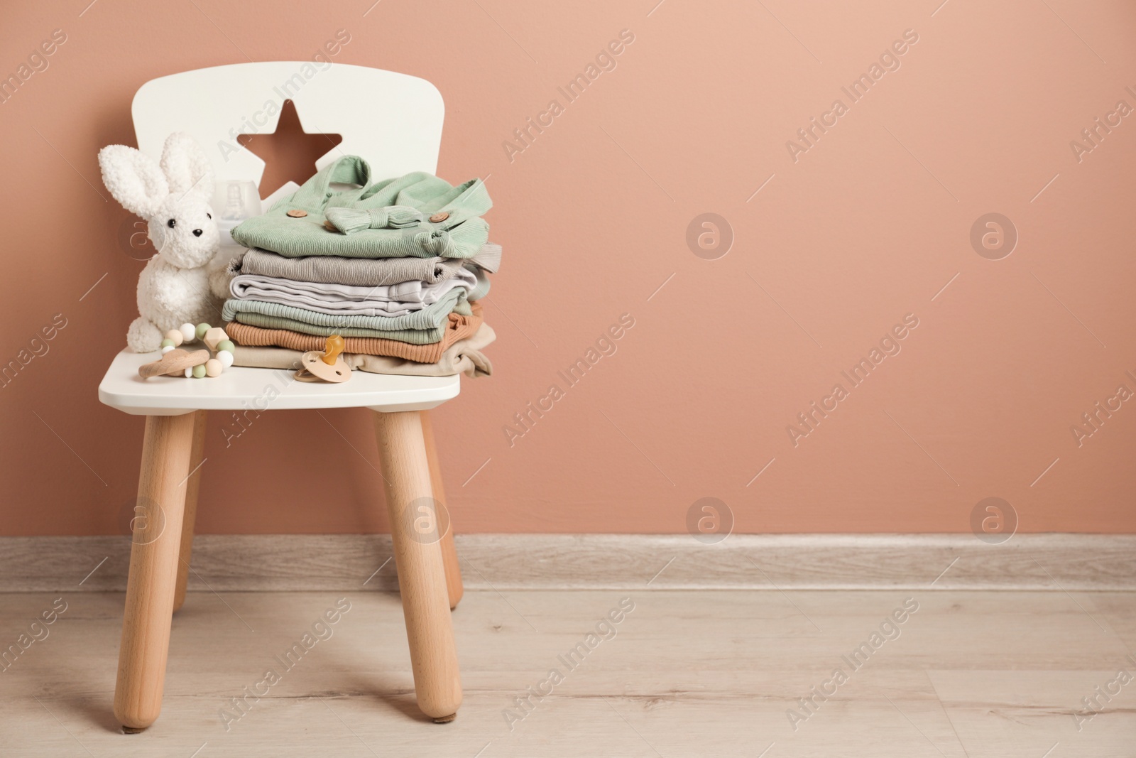 Photo of Stack of baby clothes, pacifier and toys on chair near light brown wall. Space for text