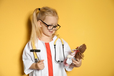 Photo of Cute child imagining herself as doctor while playing with reflex hammer and doll on color background