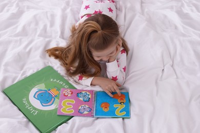 Photo of Cute little girl reading book on bed at home, above view