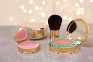 Face bronzer, blusher and other cosmetic products on grey textured table against blurred lights, closeup