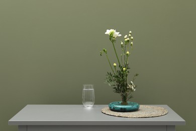 Photo of Stylish ikebana with beautiful flowers, green branches and glass of water on gray nightstand near olive wall, space for text