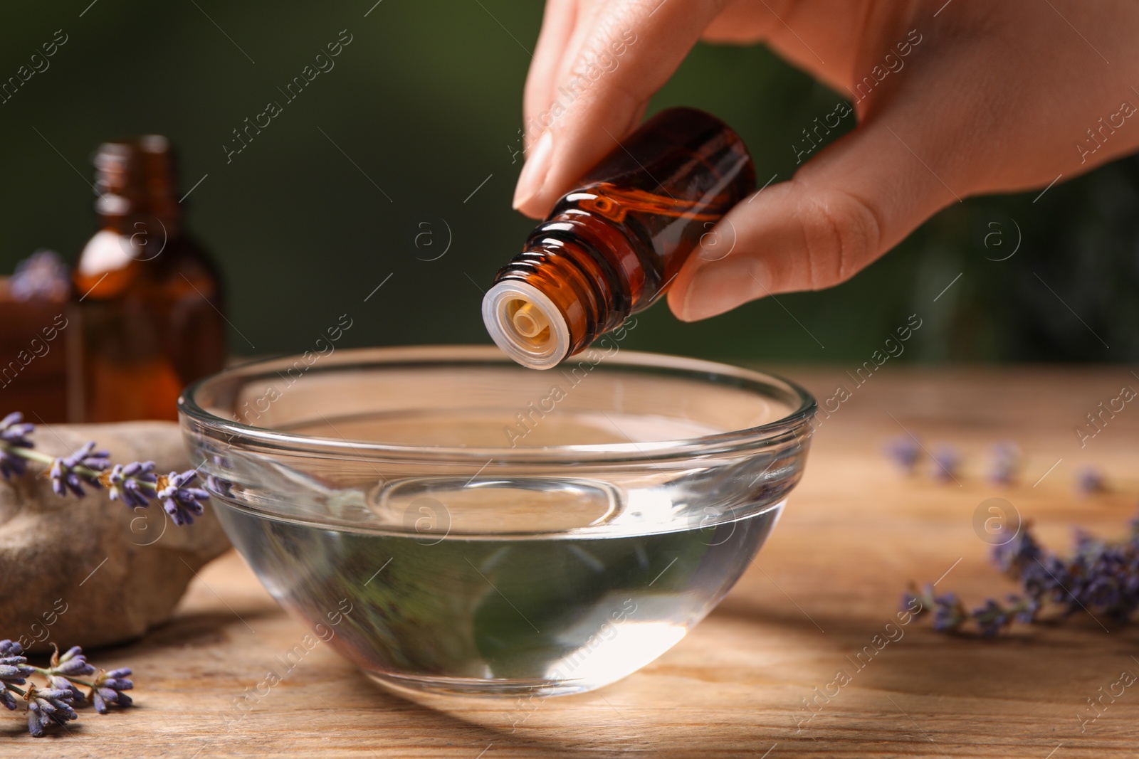 Photo of Woman dripping lavender essential oil from bottle into bowl at wooden table, closeup