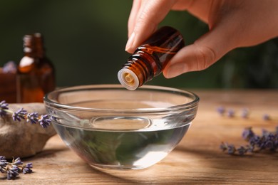 Woman dripping lavender essential oil from bottle into bowl at wooden table, closeup