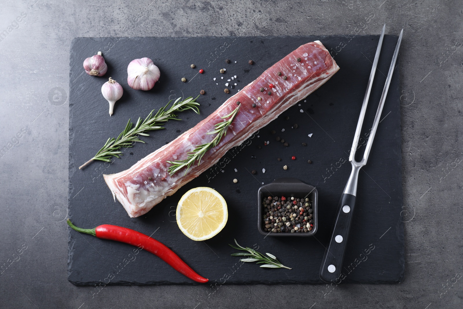 Photo of Piece of raw pork belly, rosemary, garlic, lemon and spices on grey textured table, top view