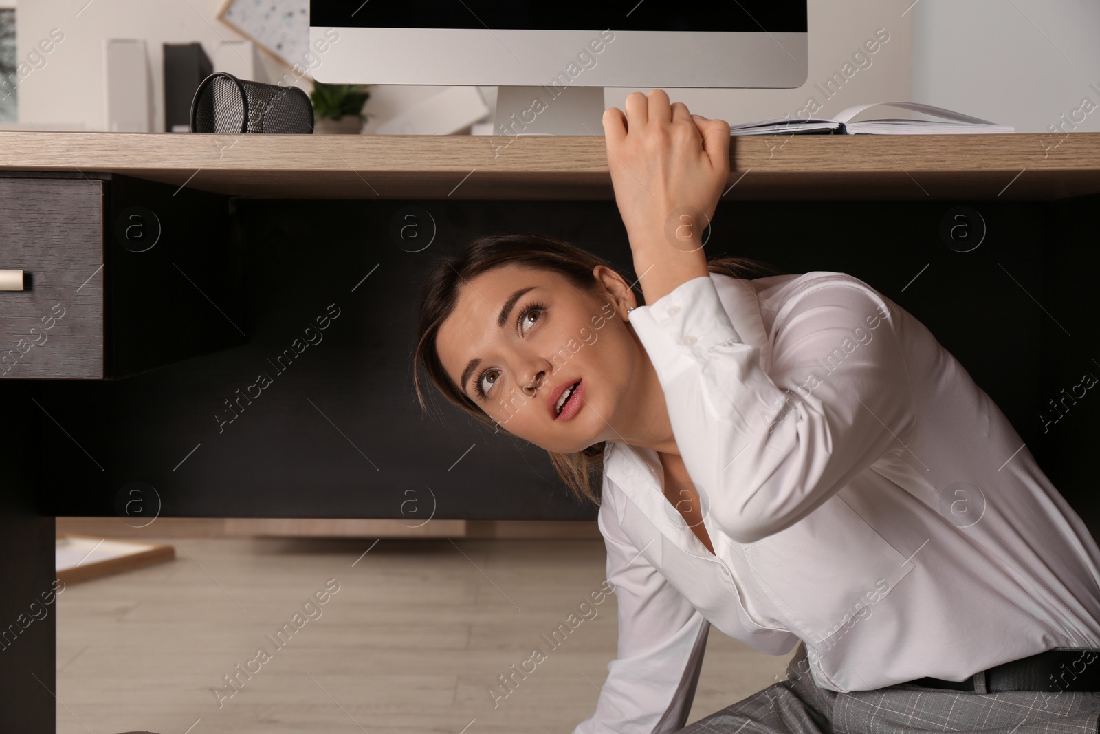 Photo of Scared young woman hiding under office desk during earthquake