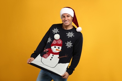 Photo of Happy man in Santa hat showing his Christmas sweater on yellow background